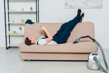 happy cleaner using smartphone while resting on sofa near vacuum cleaner  clipart
