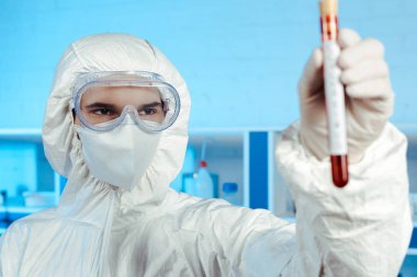 Koronavirüs harfleriyle test tüpü tutan tehlikeli madde kostümlü ve gözlüklü bilim adamlarının seçici odak noktası. 