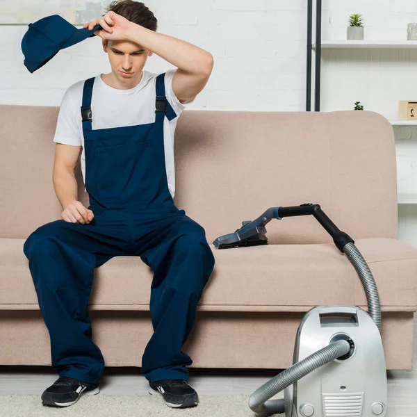 Tired Cleaner Overalls Holding Cap While Sitting Sofa Vacuum Cleaner — Stock Photo, Image