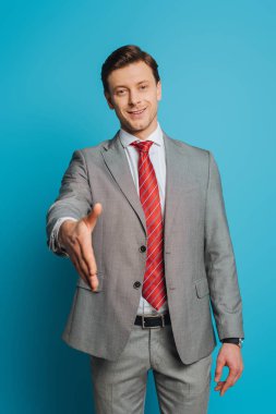 Gülümseyen iş adamı, mavi arkaplandaki kameraya bakarken elini uzatarak selam veriyor.