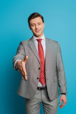 smiling businessman showing greeting gesture with outstretched hand while looking at camera on blue background clipart