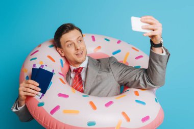 Yüzme ringinde mutlu bir iş adamı mavi arka planda pasaportları ve uçak biletlerini tutarken selfie çekiyor.