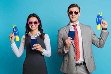 two happy businesspeople in sunglasses holding water guns, passports and air tickets on blue background clipart