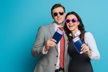 Güneş gözlüklü mutlu işadamı ve iş kadını ellerinde pasaportları ve mavi arka planda uçak biletleri var.