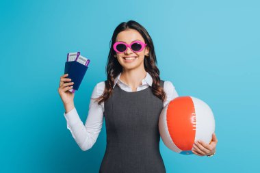 Güneş gözlüklü gülümseyen iş kadını elinde şişirilebilir top, pasaport ve mavi arka planda uçak biletleri tutuyor.
