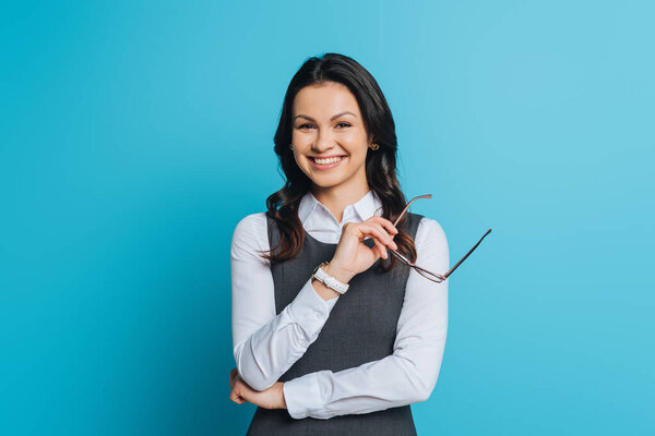 cheerful businesswoman holding eyeglasses while smiling at camera on blue background