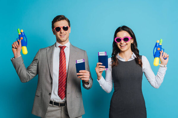 cheerful businessman and businesswoman in sunglasses holding water guns, passports and air tickets on blue background