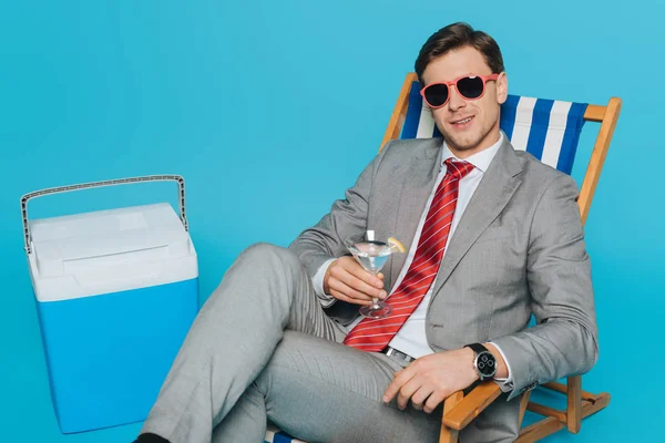 戴着太阳镜的快乐商人坐在甲板上的椅子上 在蓝色背景的便携式冰箱旁喝一杯鸡尾酒 — 图库照片