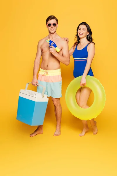 ハンサムな無修正携帯冷蔵庫とサングラスを持っています近くに笑顔ガールフレンドとともに泳ぐリングで黄色の背景 — ストック写真