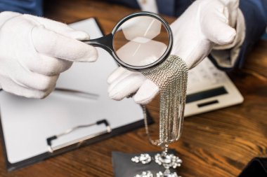 Cropped view of jewelry appraiser holding necklace and magnifying glass near calculator, clipboard and earrings on table clipart