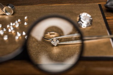 Selective focus of magnifying glass, jewelry ring with gemstone in tweezers on board on wooden table clipart