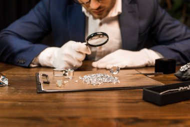 Cropped view of jewelry appraiser examining gemstones on board near jewelry on table isolated on black  clipart
