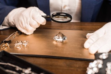 Cropped view of jewelry appraiser examining gemstone with magnifying glass near jewelry on board on table isolated on black  clipart