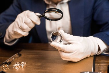 Cropped view of jewelry appraiser holding gemstone and magnifying glass near jewelry rings on board on table isolated on black clipart