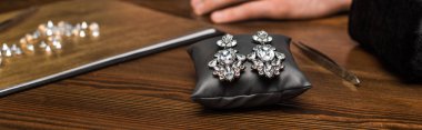 Selective focus of earrings on jewelry pillow near gemstones and hand of jewelry appraiser on wooden table, panoramic shot clipart