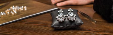 Selective focus of earrings on jewelry pillow near gemstones and hand of jewelry appraiser on wooden table, panoramic shot clipart