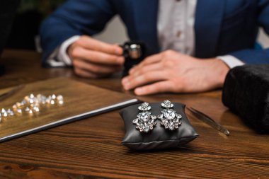 Selective focus of earrings on jewelry pillow and jewelry appraiser working at table in workshop clipart