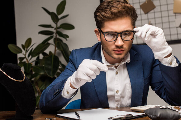 Jewelry appraiser in eyeglasses examining gemstone near calculator and clipboard on table in workshop