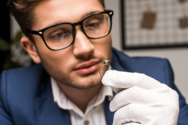 Selective focus of jewelry appraiser in glove and eyeglasses holding ring with gemstone in workshop