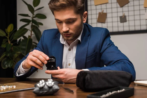 Jewelry appraiser examining jewelry ring with magnifying glass near jewelry and gemstones on table in workshop