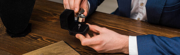 Cropped view of jewelry appraiser examining jewelry ring with magnifying glass at table on grey background, panoramic shot