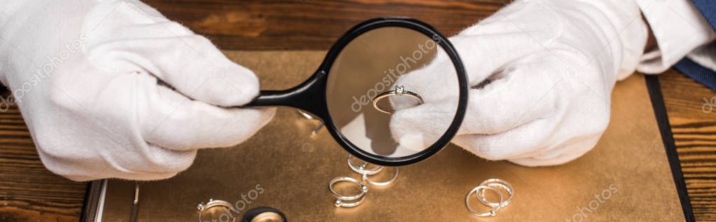 Cropped view of jewelry appraiser holding jewelry ring and magnifying glass near board on table, panoramic shot 