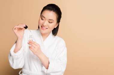 smiling beautiful asian woman in bathrobe holding dropper and bottle with serum on beige background clipart