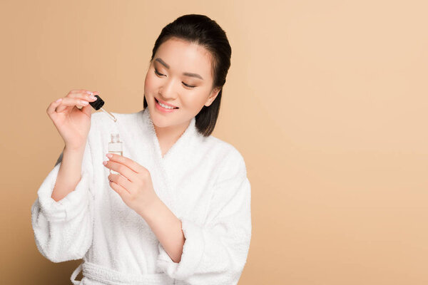smiling beautiful asian woman in bathrobe holding dropper and bottle with serum on beige background