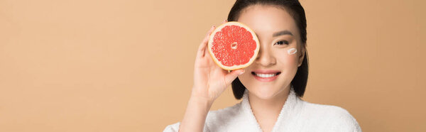 smiling beautiful asian woman in bathrobe with face cream on cheek and grapefruit half on beige background, panoramic shot