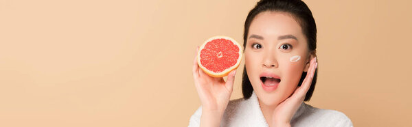shocked beautiful asian woman in bathrobe with face cream on cheek and grapefruit half on beige background, panoramic shot
