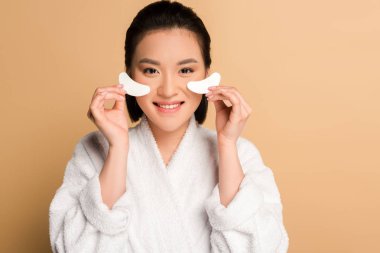 smiling beautiful asian woman in bathrobe holding eye patches on beige background clipart