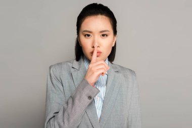 young businesswoman in suit showing shh gesture on grey background clipart