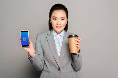 KYIV, UKRAINE -  FEBRUARY 11, 2019: happy young businesswoman with paper cup holding smartphone with shazam app on grey background