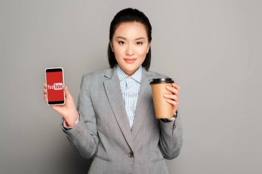 KYIV, UKRAINE -  FEBRUARY 11, 2019: happy young businesswoman with paper cup holding smartphone with youtube app on grey background