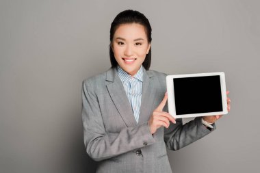 Takım elbiseli mutlu genç iş kadını gri arka planda dijital tablet tutuyor.