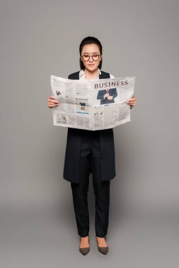 Gözlüklü genç iş kadını gri arka planda gazete okuyor.