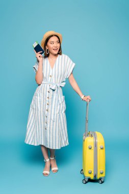 Sırıtan esmer Asyalı kız çizgili elbise ve hasır şapkalı seyahat çantası ve mavi arka planda uçak bileti olan pasaport