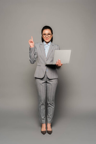 улыбающаяся молодая деловая женщина в очках с ноутбуком и показывающая жест идеи на сером фоне
