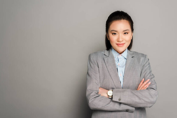 smiling young businesswoman in suit with crossed arms on grey background