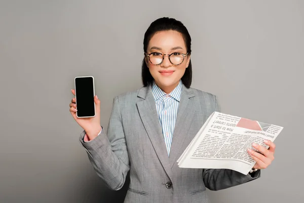 眼鏡をかけて新聞やスマートフォンを灰色の背景に持つ若い実業家が — ストック写真