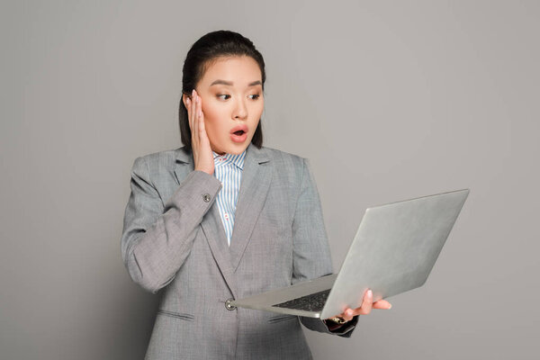 шокированная молодая деловая женщина в костюме с ноутбуком на сером фоне
