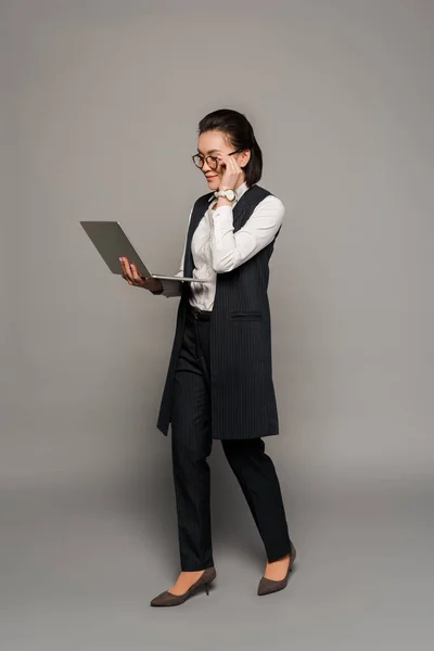 戴眼镜的年轻女商人在灰色背景下使用笔记本电脑 — 图库照片