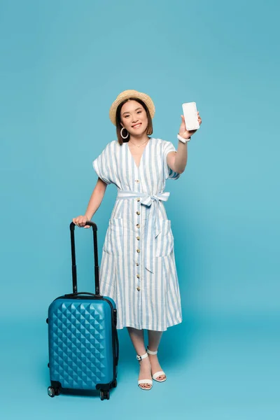 笑顔ブルネットアジアの女の子でストライプドレスとストロー帽子とともに旅行バッグとスマートフォン上の青の背景 — ストック写真