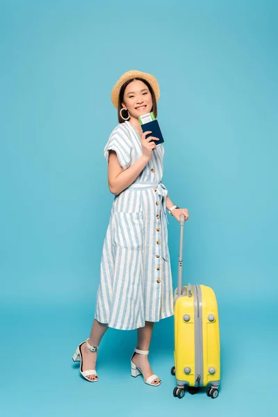 笑顔ブルネットアジアの女の子でストライプドレスとストロー帽子とともに旅行バッグとパスポート付き航空券で青の背景 — ストック写真
