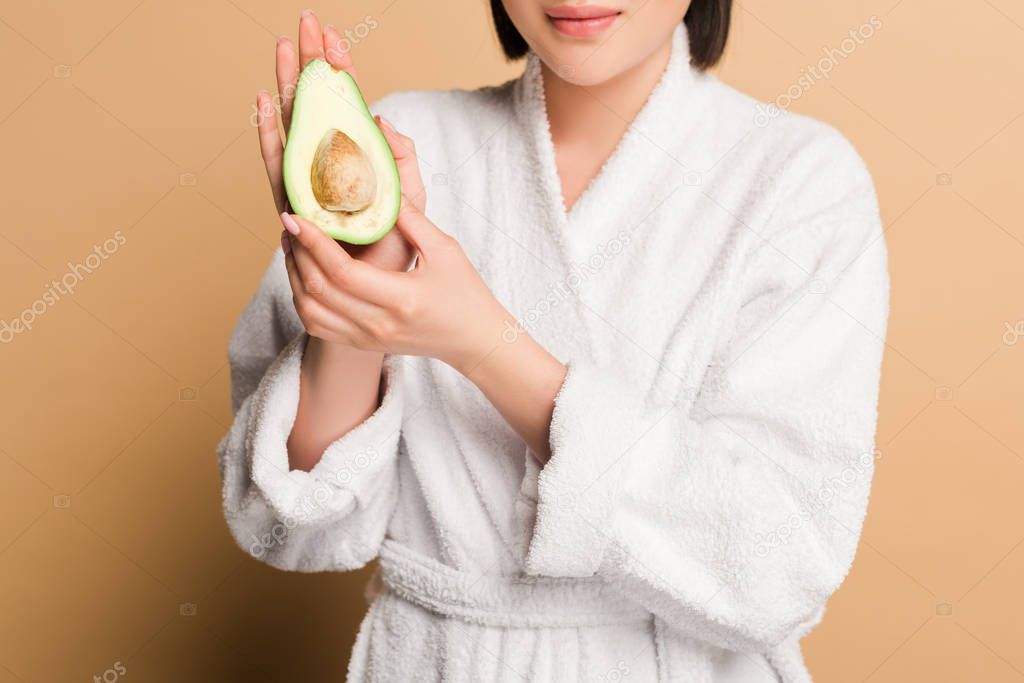 cropped view of happy beautiful asian woman in bathrobe with avocado half on beige background