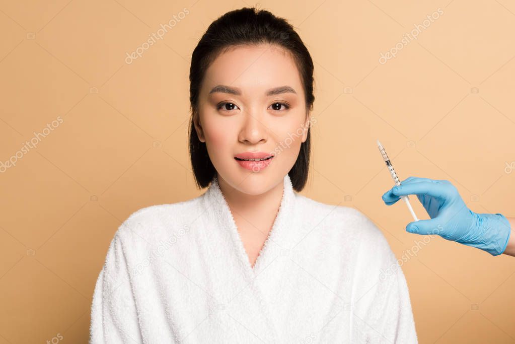 beautiful asian woman in bathrobe near cosmetologist hand with syringe for beauty injection on beige background