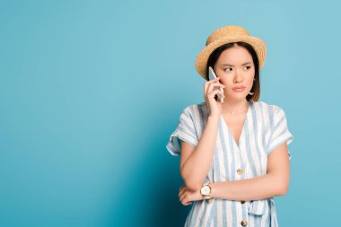 üzgün esmer Asyalı kız çizgili elbiseli ve hasır şapkalı mavi arka planda akıllı telefonu alıyor.