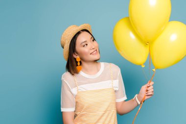 happy asian girl in striped dress and straw hat with yellow balloons on blue background clipart