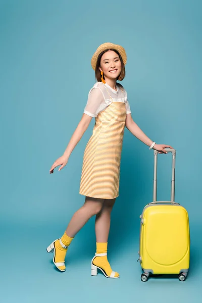 笑着的亚洲女孩 身穿条纹黄色衣服 头戴草帽 蓝色背景的手提箱 — 图库照片