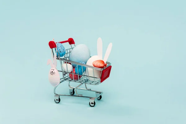 Bunte Ostereier Und Dekorative Hasen Einkaufswagen Auf Blauem Hintergrund — Stockfoto